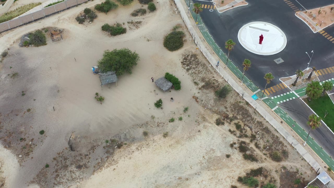 Aerial Los Cabos frame with open sand across the center, scrub vegetation, a small shade structure, the green bike lane, and a white circular paved feature at upper right.
