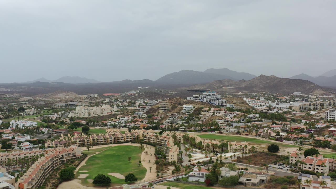 Second frame looking across Los Cabos, Mexico with a curved green fairway in the foreground, clustered residences in the midground, hazy mountains beyond, and no visible people.
