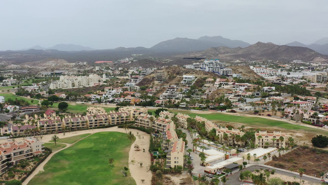 Elevated Los Cabos view showing a curved green landscaped area, rows of beige buildings, scattered white houses, dry hills, distant mountains, and no visible people.