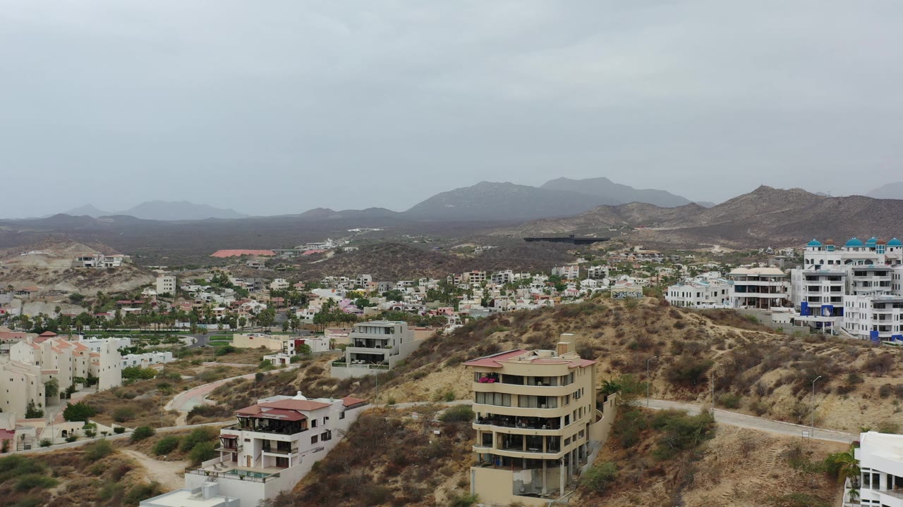 Los Cabos residential hills fill the frame with a tan multi-story house in front, white buildings beyond, mountains in the distance, and no visible people.