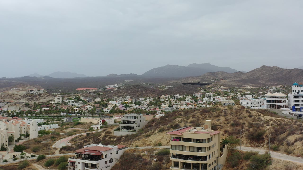 Opening aerial frame over Los Cabos, Mexico with sandy hillside homes, low-rise condos, distant mountains, overcast sky, and no visible people.