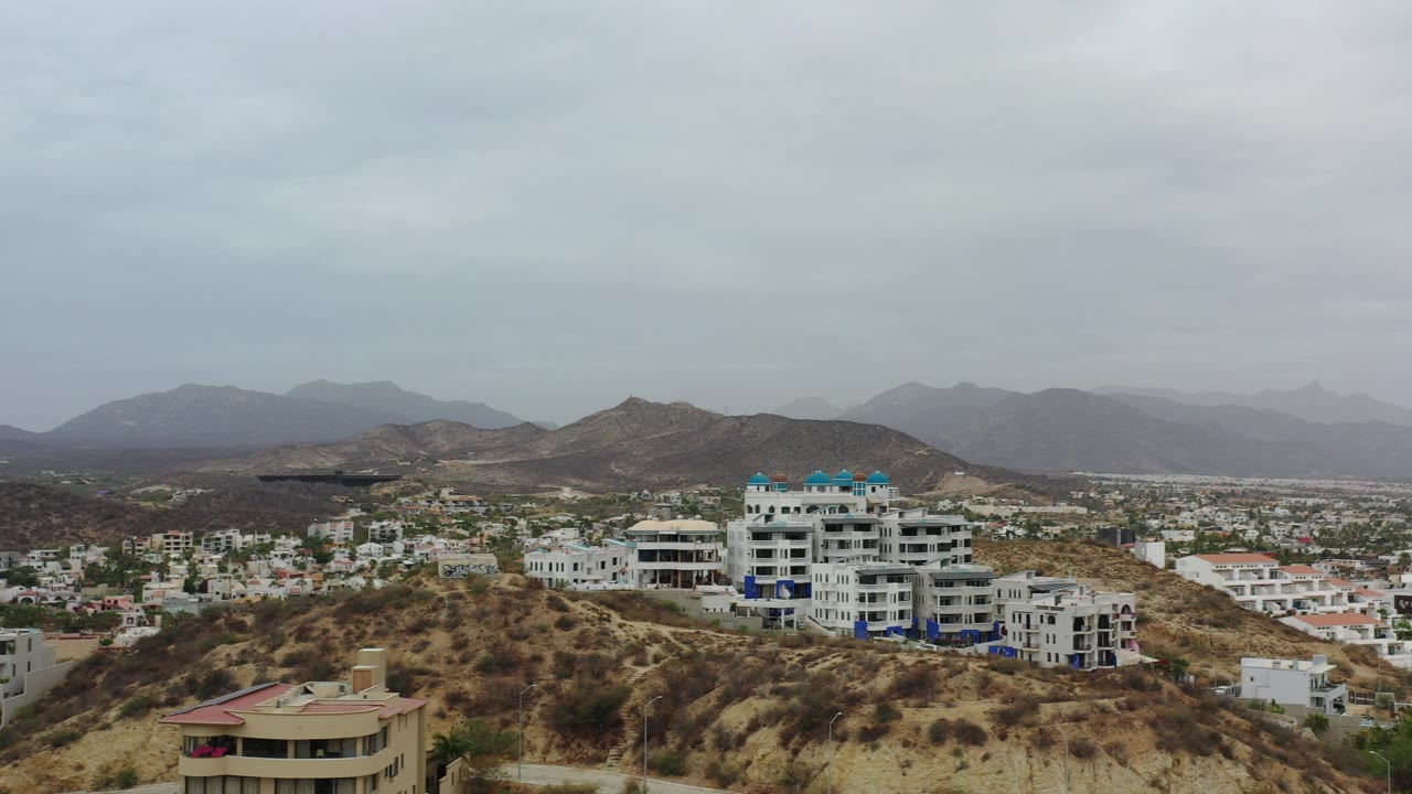 Opening frame of Los Cabos, Mexico, showing dry hills, white low-rise buildings, distant mountains, and gray cloud cover with no visible people.