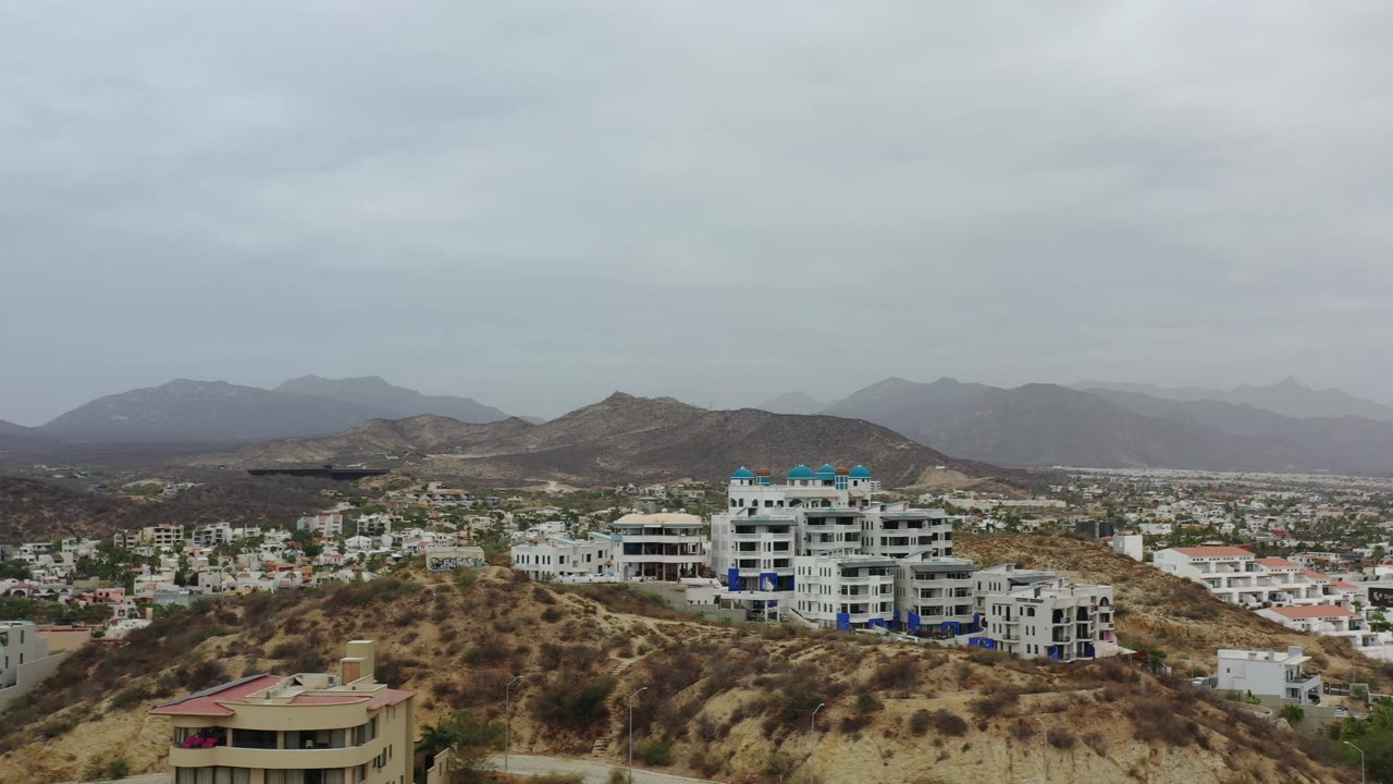 Los Cabos hillside neighborhood seen from above, with a blue-roofed white building near center, brown slopes, hazy mountains, and no visible people.