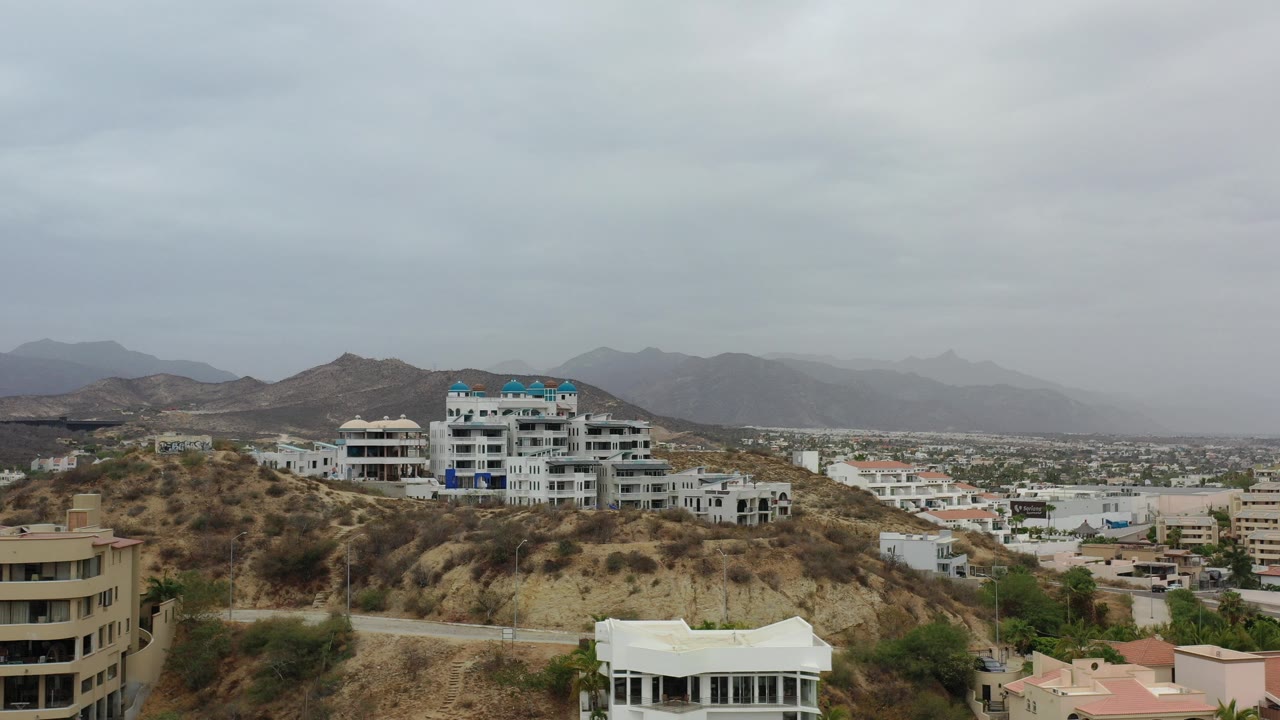 Wide hillside view with modern buildings and distant mountains under gray sky.