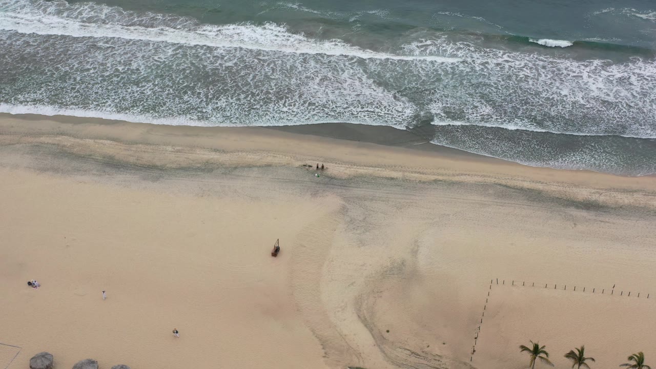 Aerial view of foamy waves pushing farther onto the wet sand in Los Cabos, Mexico, with small beach objects below and no visible people or faces.