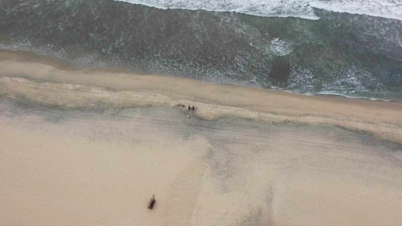 Overhead view of the Los Cabos shoreline where dark ocean water and white foam meet tan sand, with no visible people.