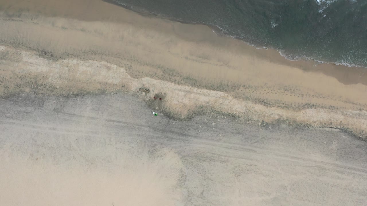 Top-down view of a Los Cabos beach with dark surf at the top, pale sand below, and tiny people or beach items near the shoreline.