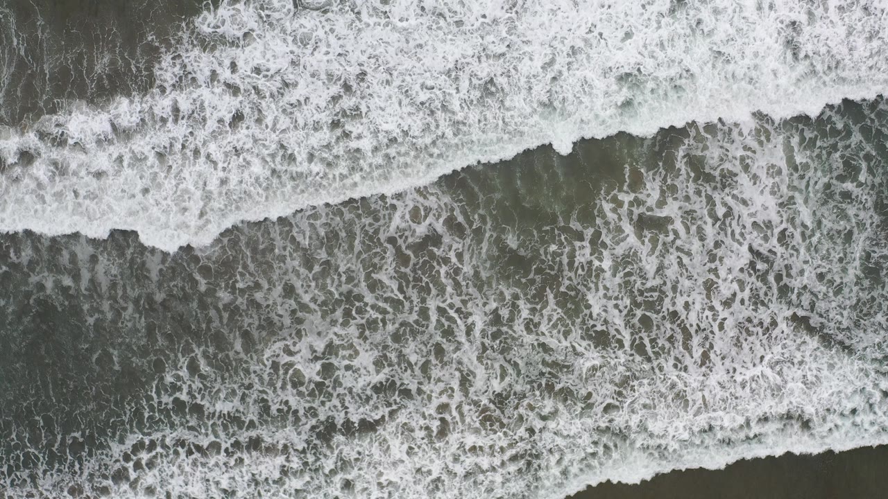 Overhead view of white surf foam spreading across darker shallow water in Los Cabos, Mexico, with no visible people.