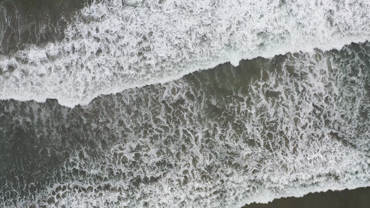 Aerial top-down frame of broken white foam over grey-green shallow ocean in Los Cabos, Mexico, with no visible people.