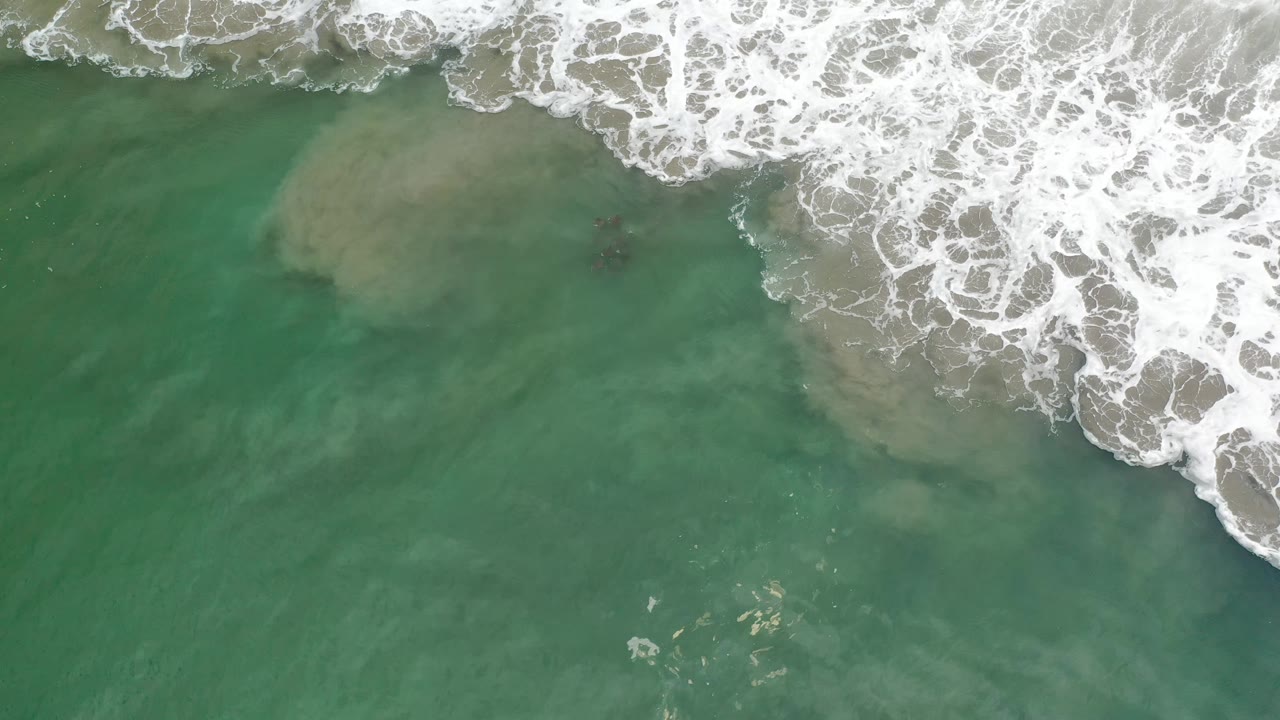 Aerial view in Los Cabos, Mexico, with white foam pushing in from the upper right over green water and no visible people.