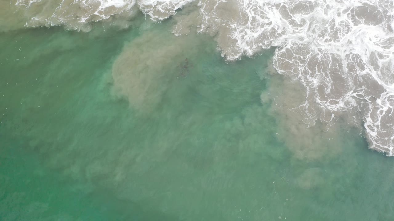 Aerial frame of white foam curling across aqua shallow water and pale sand in Los Cabos, Mexico, with no visible people.