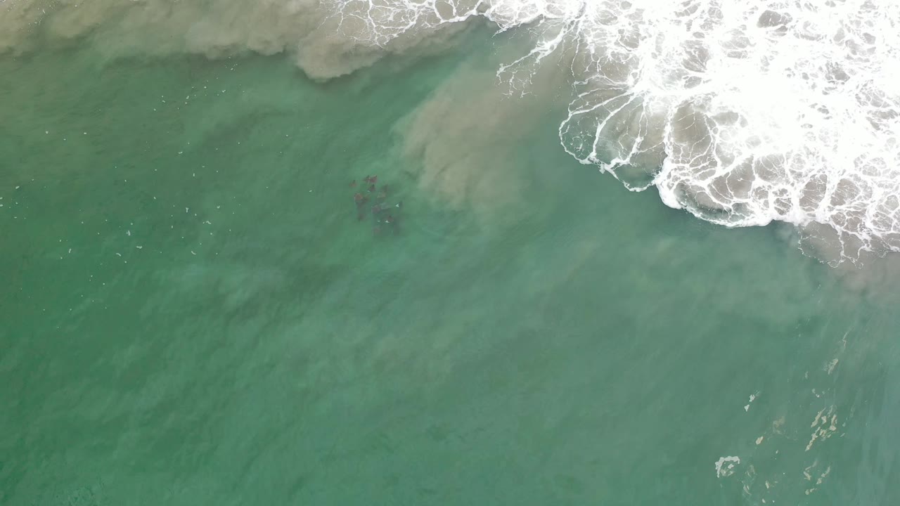 A top-down Los Cabos ocean frame shows foamy surf spreading farther across green water, with a faint dark shape under the surface and no visible people.