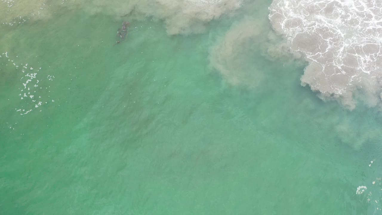 Aerial frame over Los Cabos water, showing milky sand movement and white foam near the upper border with no visible people.