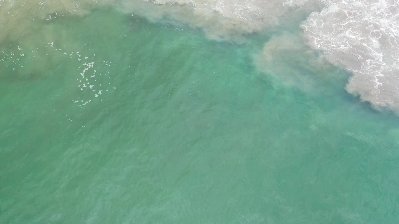 Overhead view of turquoise ocean water in Los Cabos, Mexico, with white surf foam entering along the top edge and no visible people.