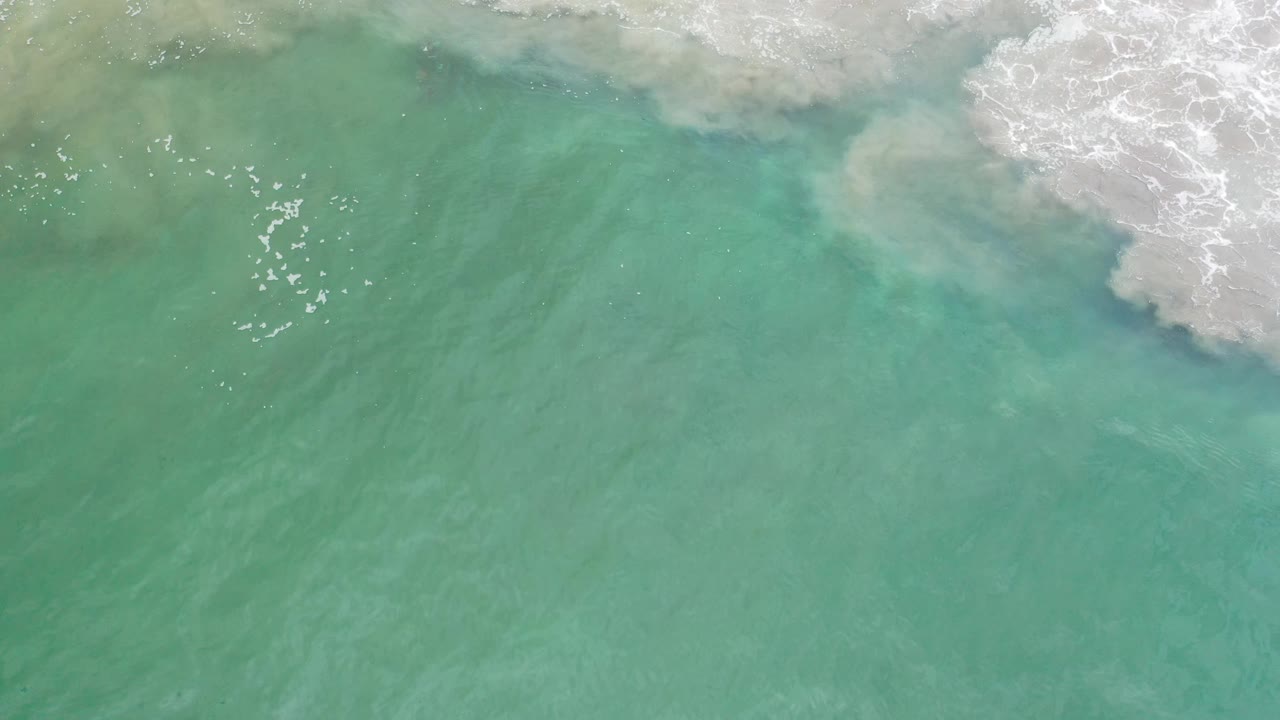 Overhead Los Cabos ocean frame with blue-green water, pale sand-toned swirls under the surface, white wave wash near the top, and no visible people.