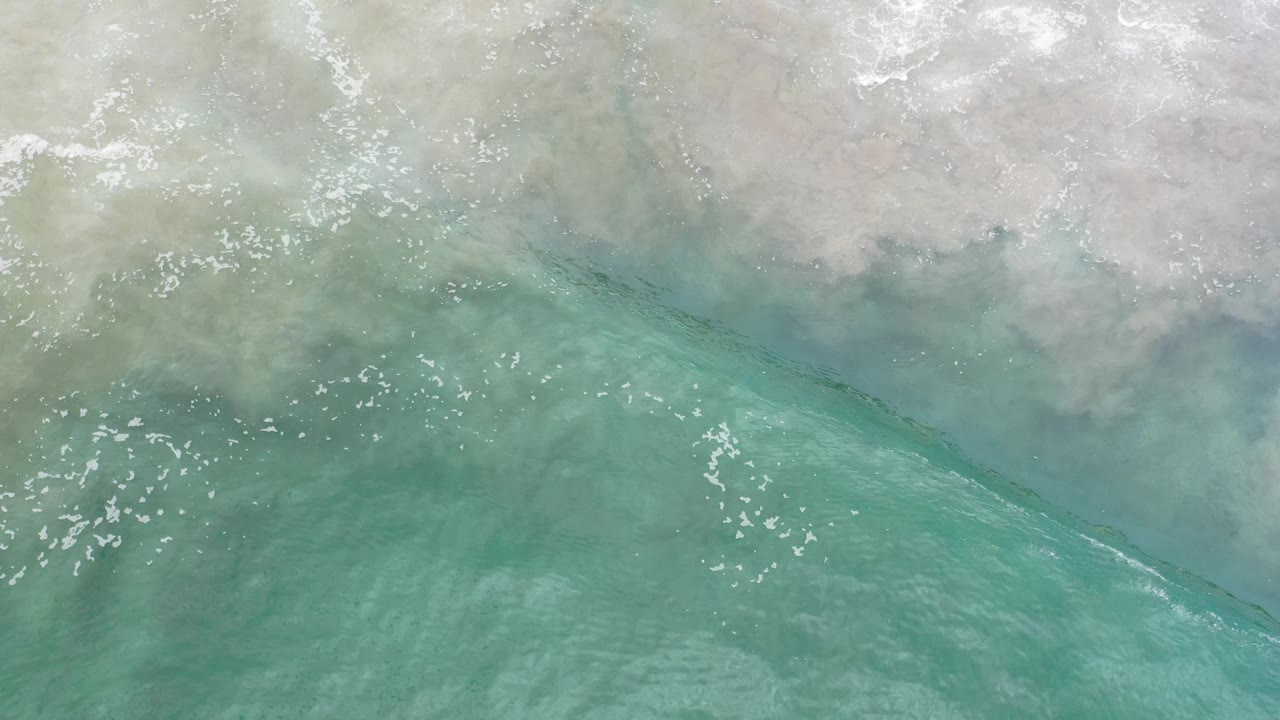 Aerial frame from Los Cabos, Mexico, showing blue-green water curling diagonally beside pale sand with scattered white highlights, no visible people.