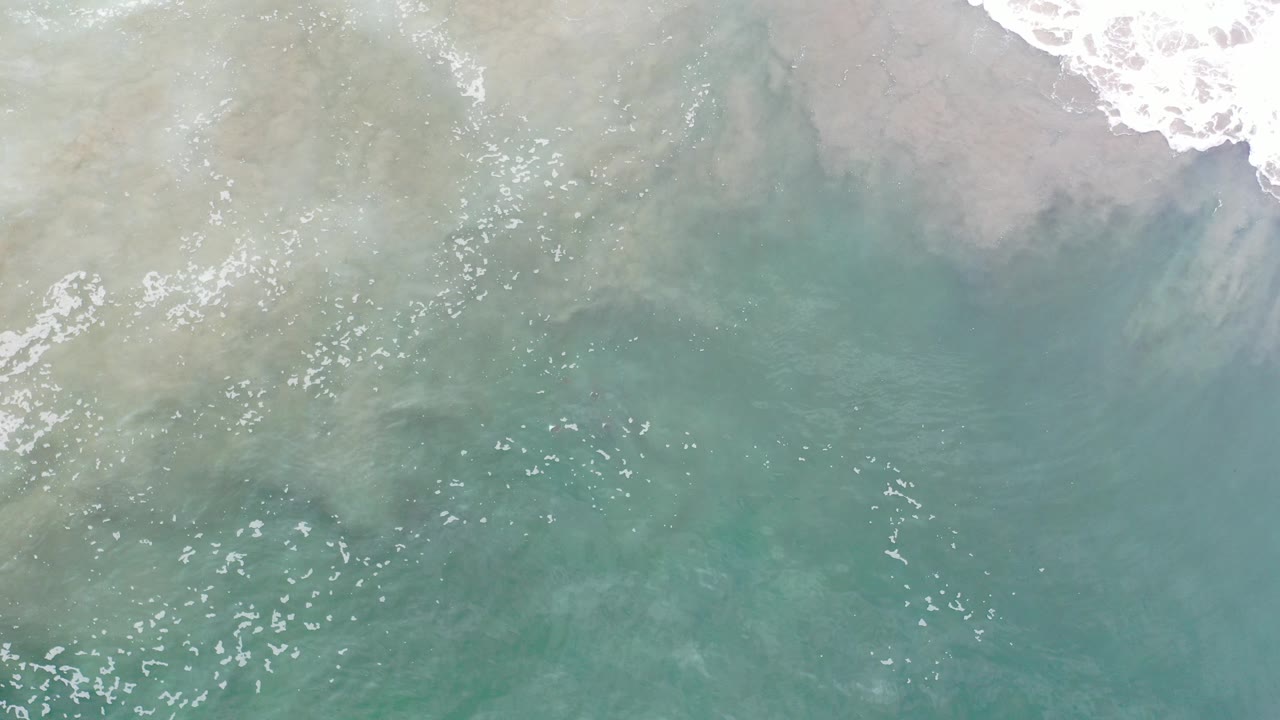 Overhead view of pale turquoise water in Los Cabos, Mexico, with white surf foam entering along the upper edge and no visible people.