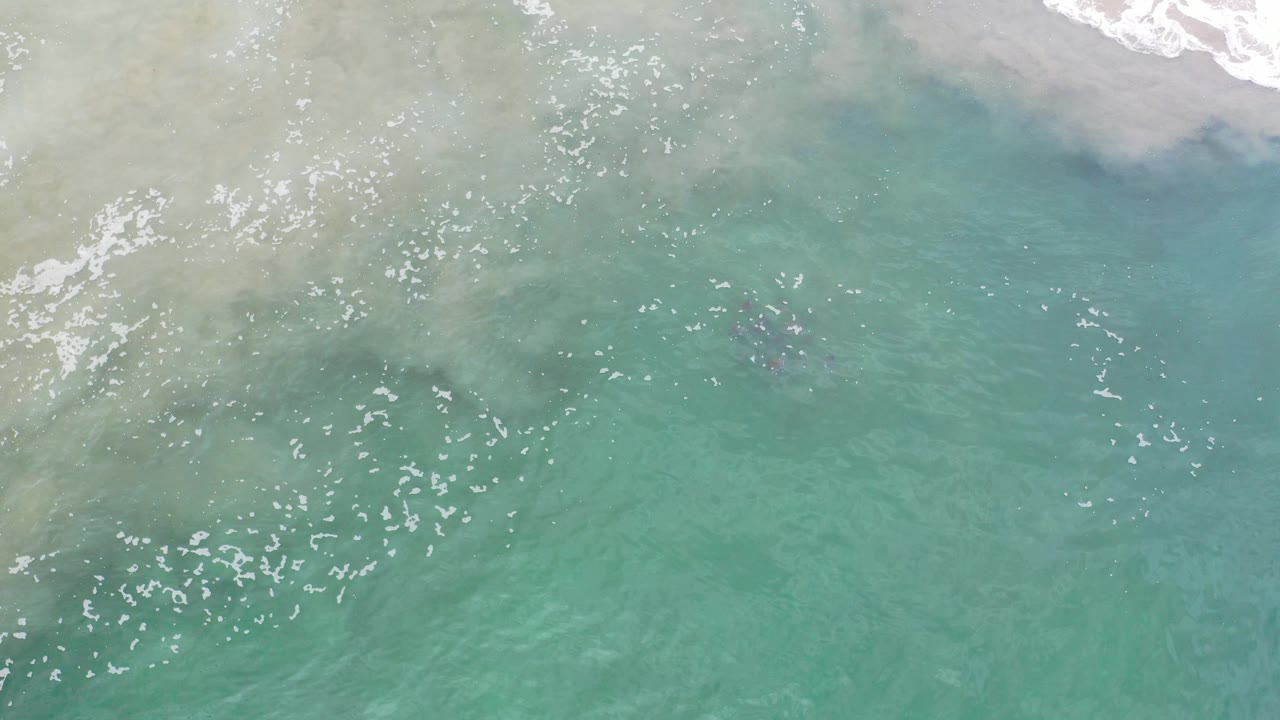 Opening overhead frame from Los Cabos, Mexico, showing pale foam along the upper edge, clear green-blue shallow water, faint darker underwater shapes, and no visible people.
