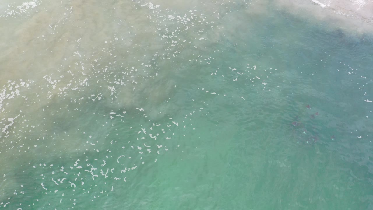 Overhead view of turquoise ocean water in Los Cabos, Mexico, with pale sand-colored clouding near the top of frame, scattered sun reflections, faint dark marks below the surface, and no visible people.