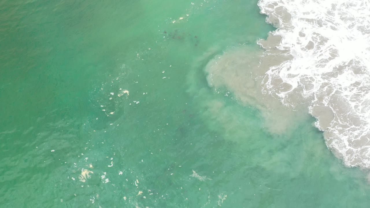 A pale sandy patch shows through green-blue water in Los Cabos, Mexico, while broken white foam gathers along the upper right with no visible people.