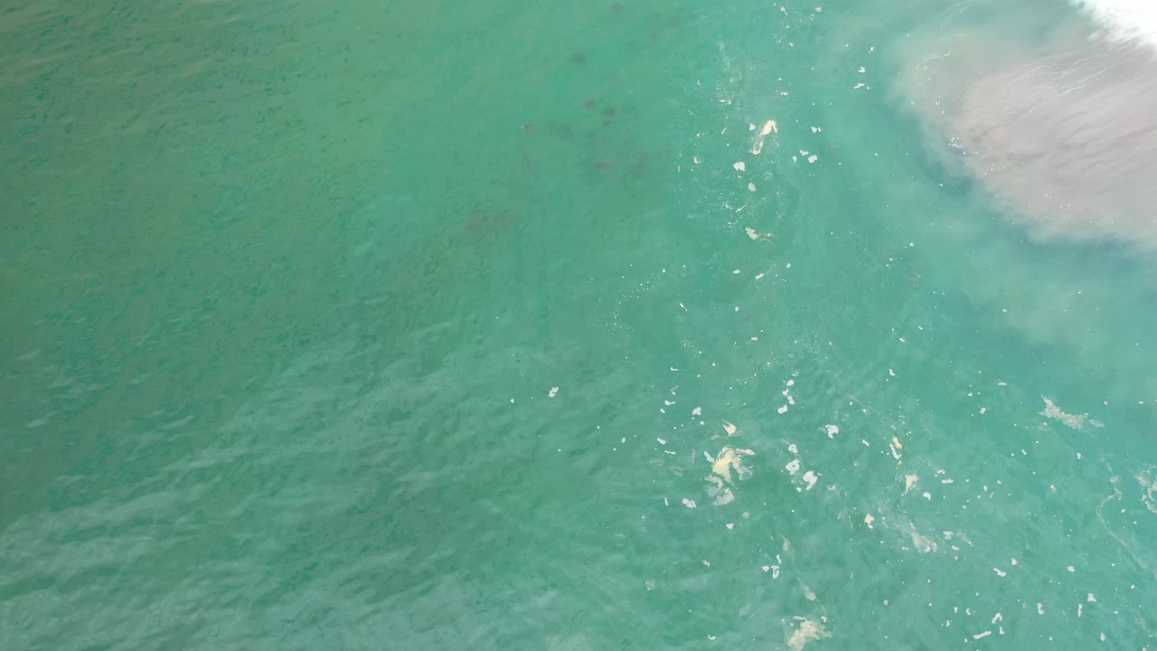 Aerial-style view of Los Cabos green water with small surface ripples, scattered highlights, and a pale sand or foam patch entering the upper right, with no visible people.