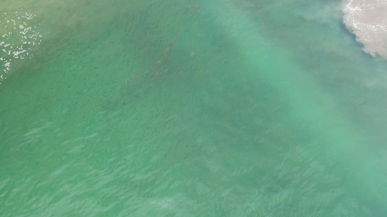 Overhead view of turquoise-green seawater in Los Cabos, Mexico, with a white foamy patch near the top edge, faint dark submerged shapes, and no visible people.