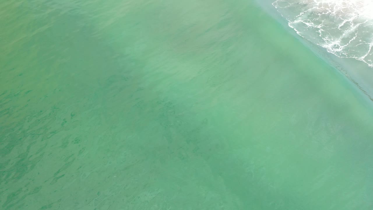 Overhead view of pale green ocean water in Los Cabos, Mexico, with a diagonal band of white surf near the top edge and no visible people.