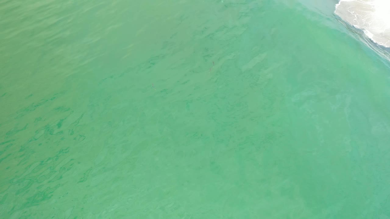 A high-angle view over Los Cabos water shows pale green surf patterns, darker streaks below the surface, and white foam along the top right, with no visible people.