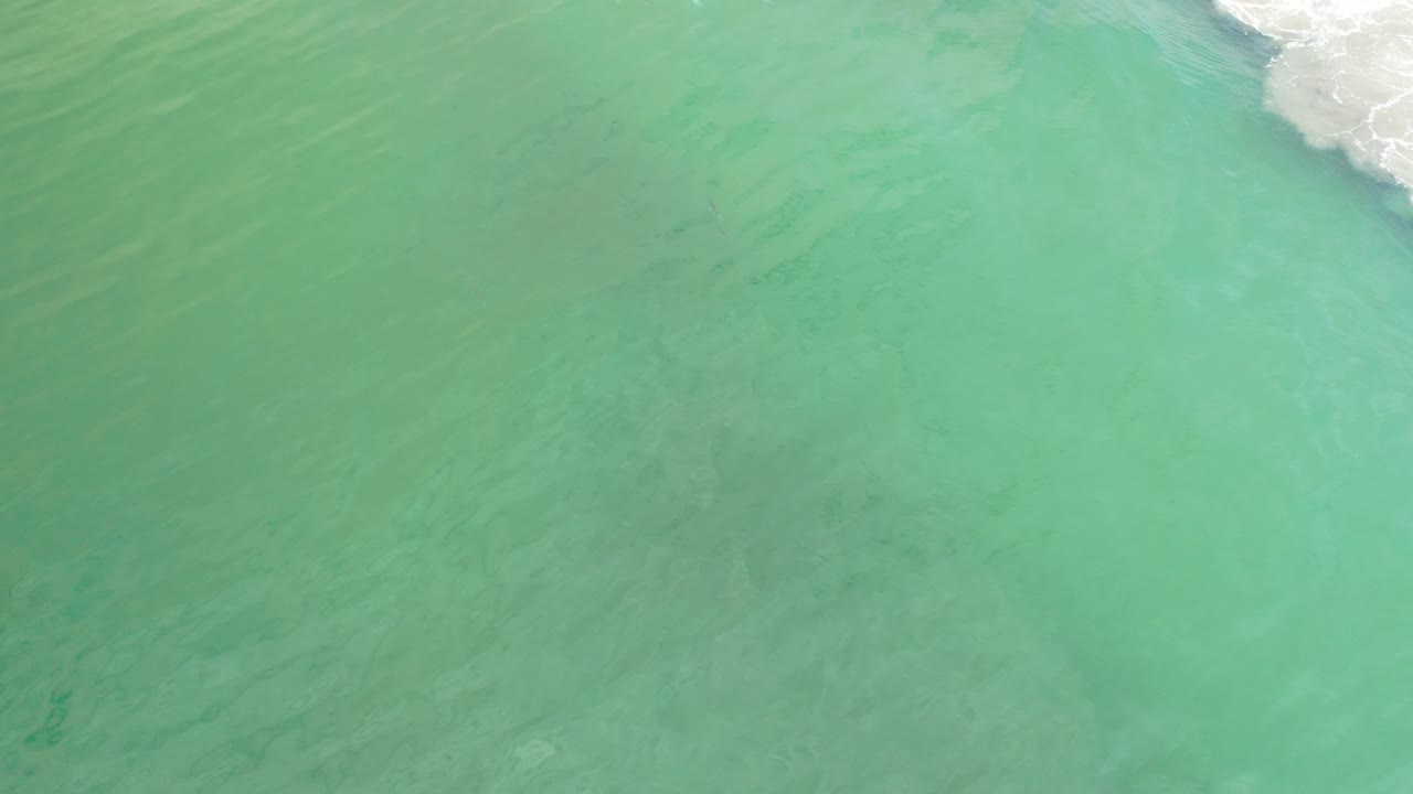 Overhead view of turquoise ocean water in Los Cabos, Mexico, with faint darker streaks below the surface and a small white wave edge at the upper right, with no visible people.