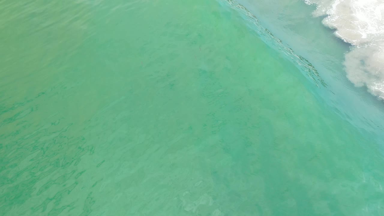 Overhead Los Cabos water frame with pale green blue shallows and breaking white foam along the top, with no visible people.