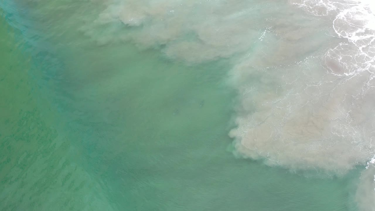 Green ocean surface with white foam and a pale underwater sandbar on the right.