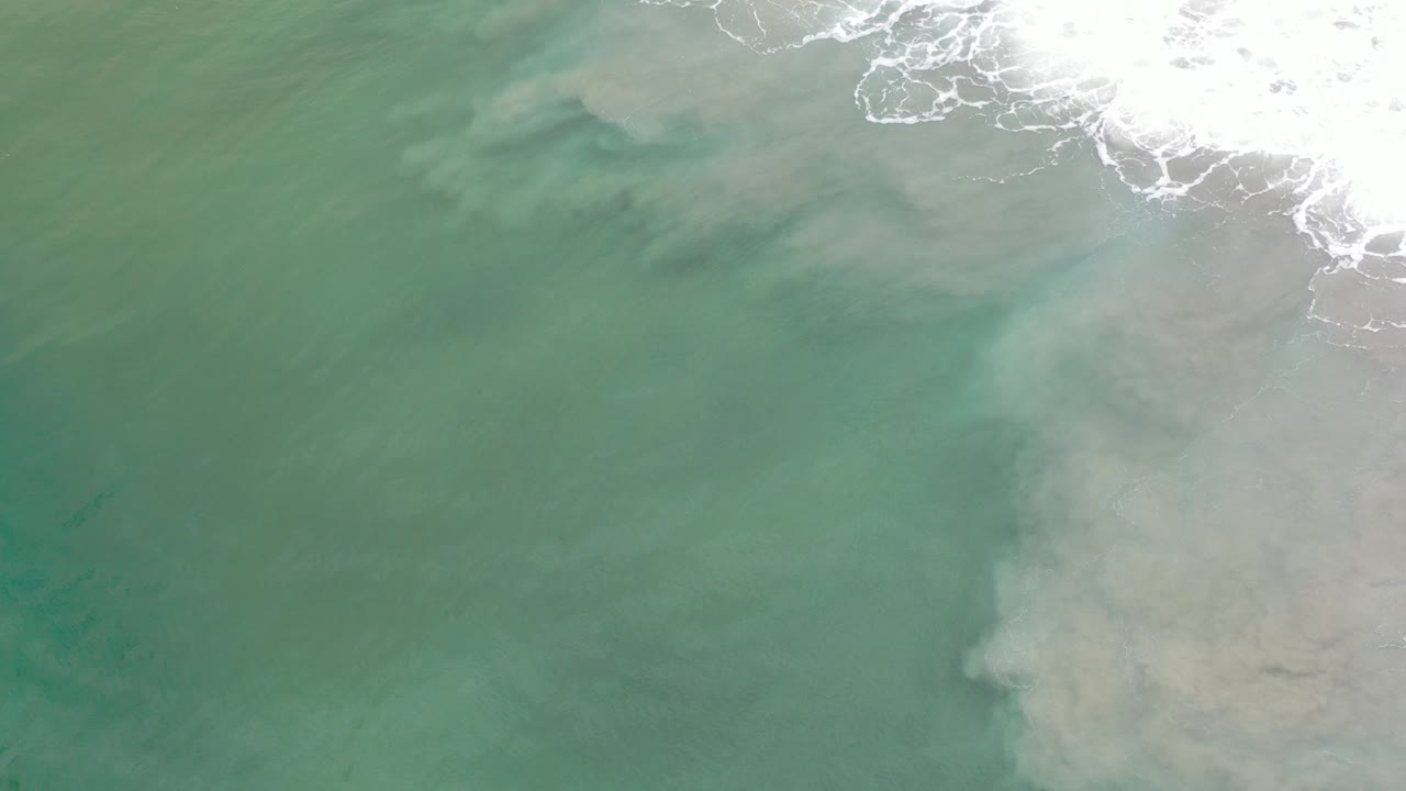 Overhead view of Los Cabos shoreline water with green-blue shallows, cloudy sand patterns below the surface, white surf along the top edge, and no visible people.
