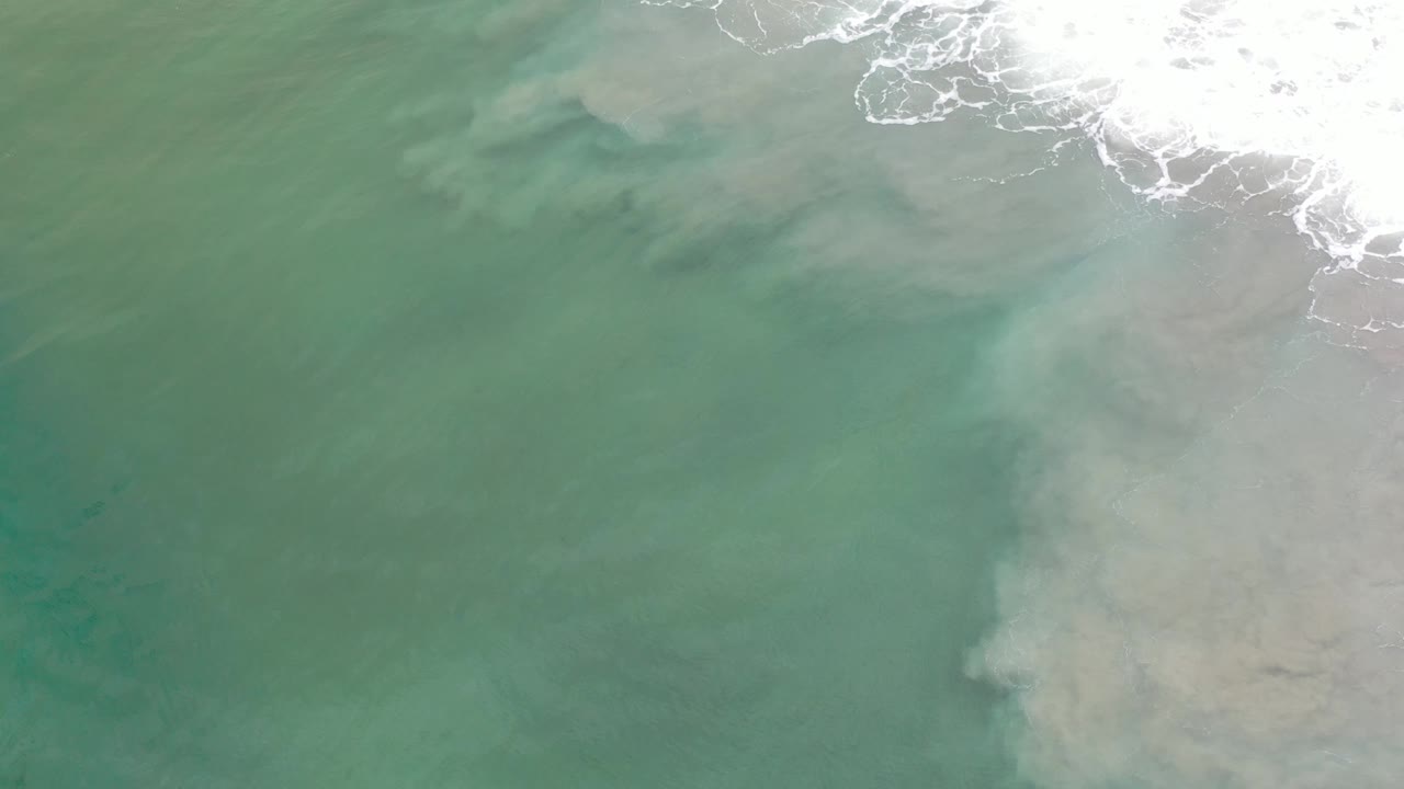 Overhead Los Cabos shoreline frame showing translucent green water, pale sandy sediment shapes beneath the surface, surf foam near the upper right, and no visible people.