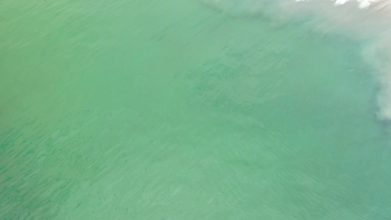 Pale green ocean water in Los Cabos fills the frame at the opening moment, with faint ripples and a small band of white foam near the top edge, no visible people.