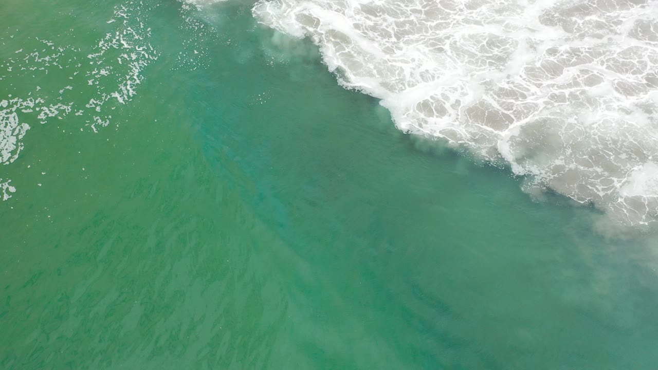 Overhead view of green-blue surf water in Los Cabos, Mexico, with white foam entering from the top edge and no visible people.
