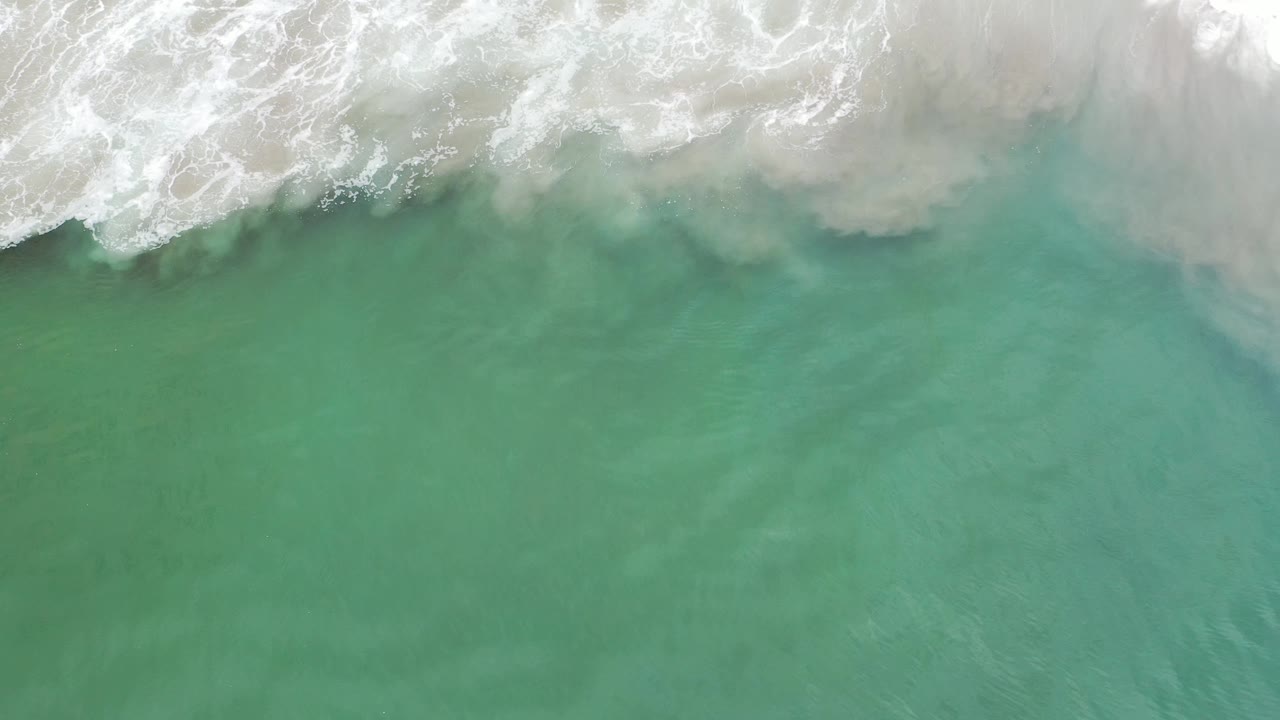 Aerial overhead frame in Los Cabos, Mexico, showing green-blue ocean below a wider strip of foamy breaking wave and no visible people.