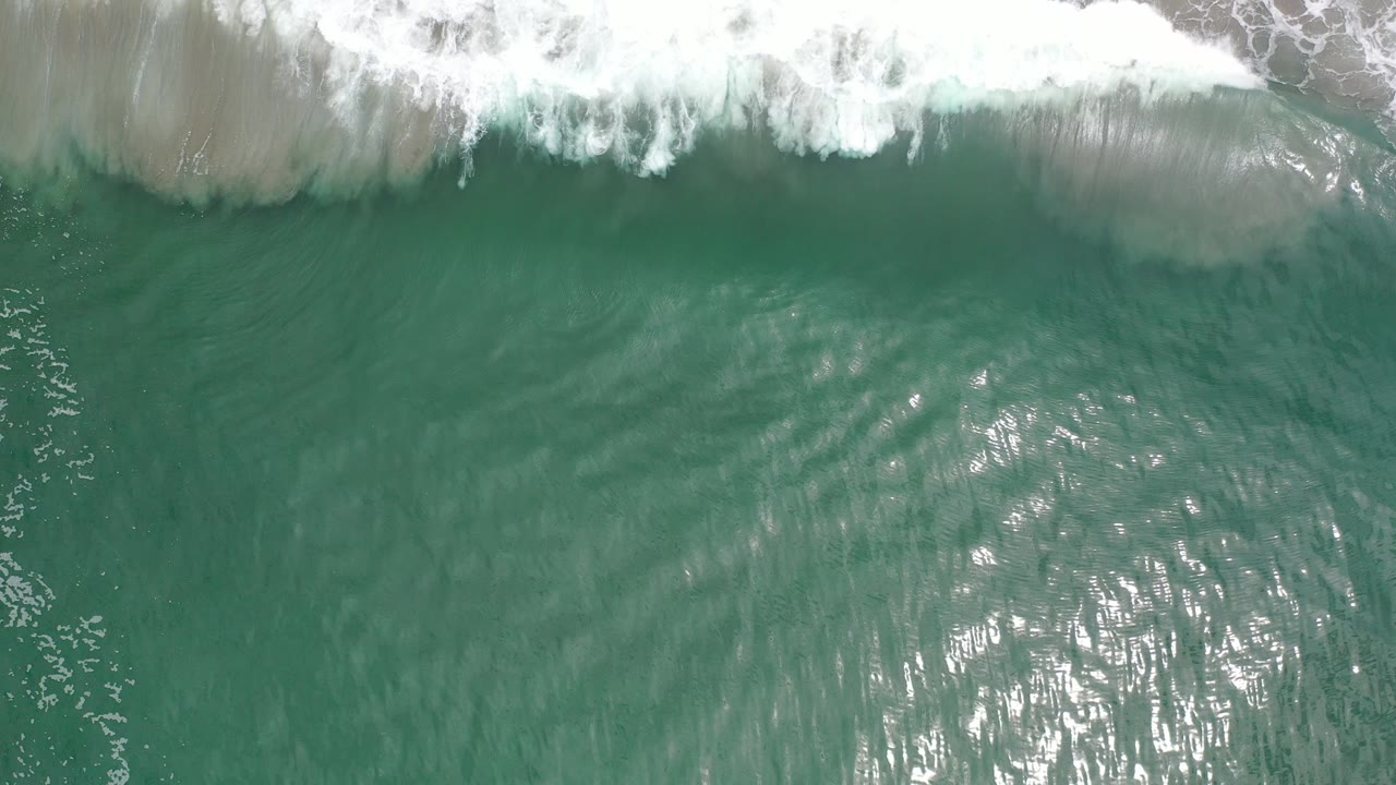 Overhead view of turquoise ocean water in Los Cabos, Mexico, with white surf breaking across the top edge, bright sun reflection below, and no visible people.