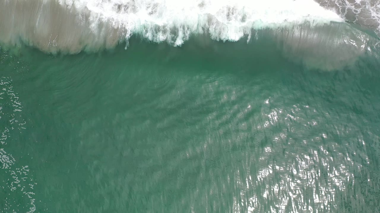 Overhead Los Cabos ocean frame with white foam still visible at the top, a bright patch of sun glare near the lower center, and no visible people.
