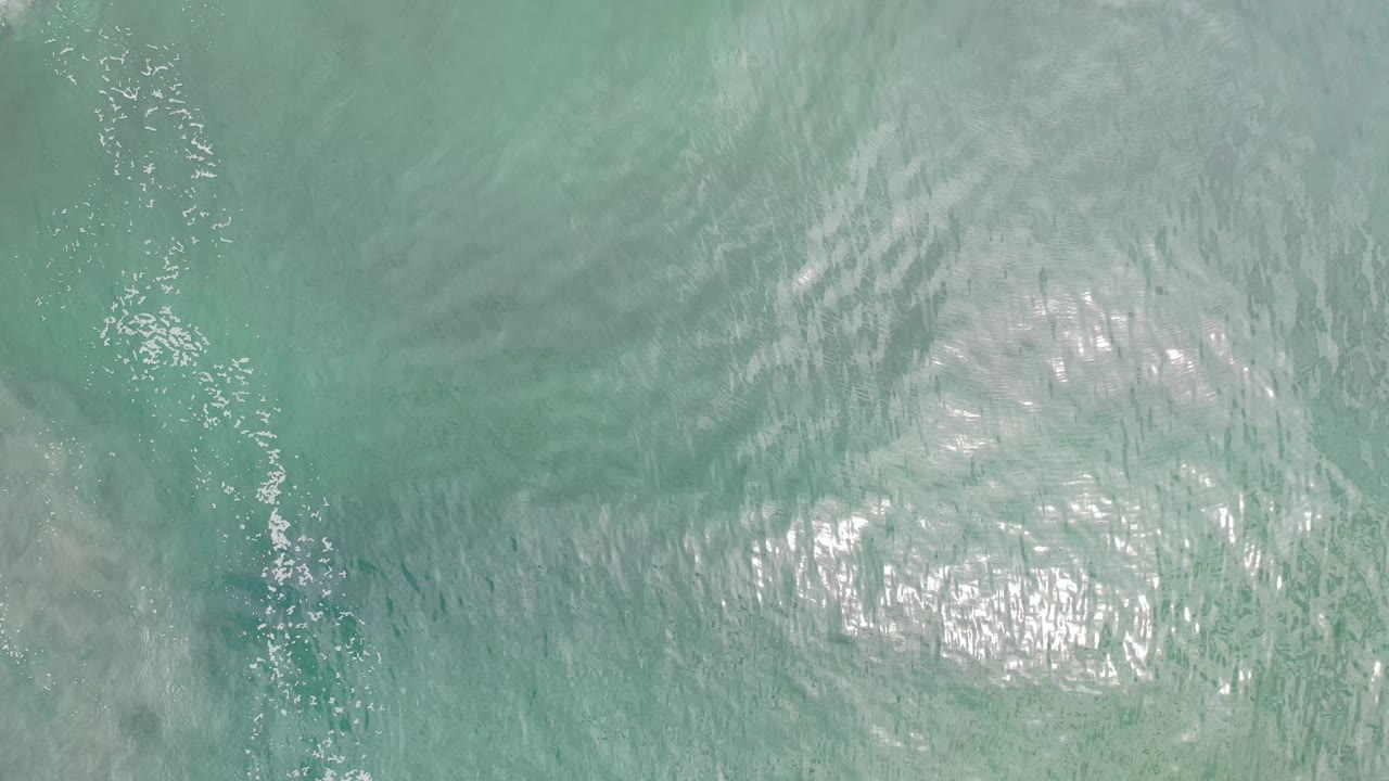 Overhead Los Cabos ocean frame showing pale green-blue water, a central patch of sunlight, faint wave texture, and no visible people.