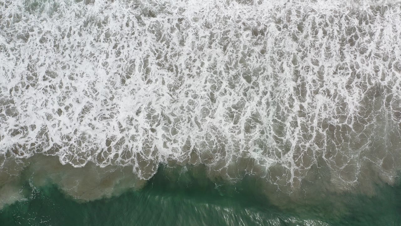 Overhead frame from Los Cabos, Mexico showing dense white foam spread across green seawater, with no visible people.