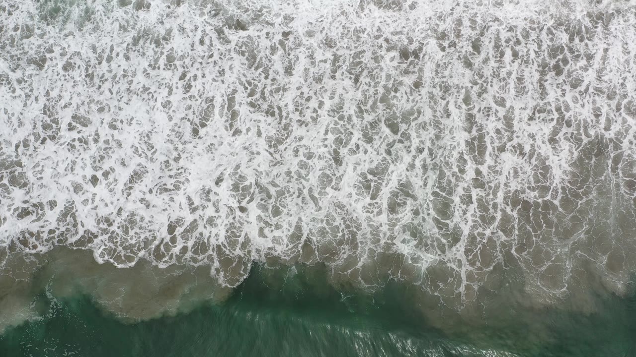 Aerial view in Los Cabos, Mexico with a thick white wave band crossing darker green water and no visible people.
