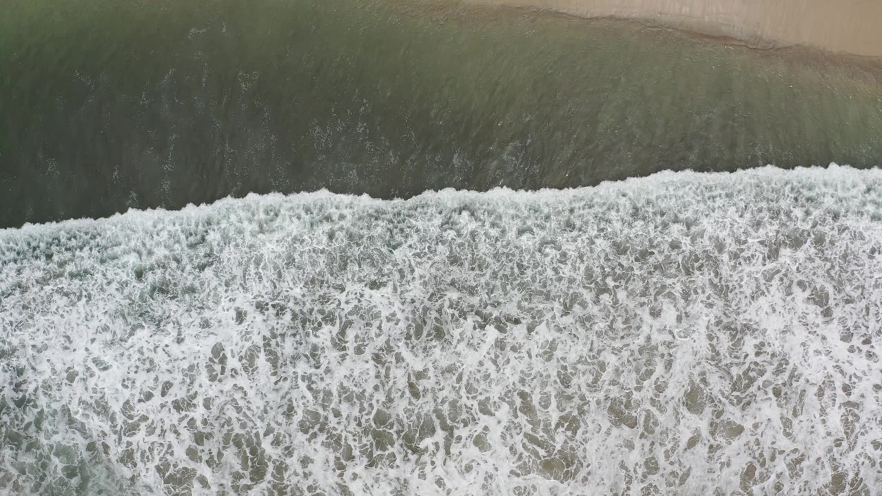 Aerial Los Cabos beach frame with a broad white foam band across the center and darker water above the sandy shore, with no visible people.