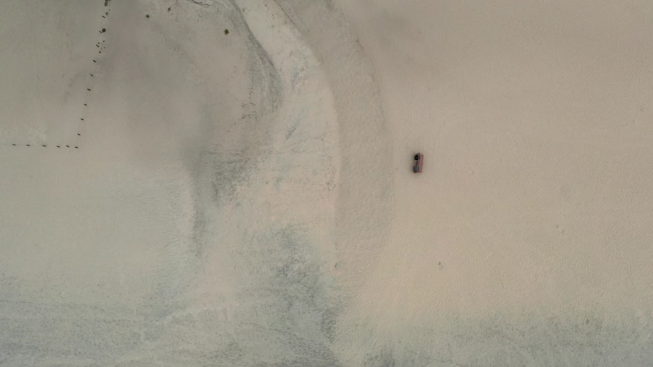 Top-down Los Cabos beach frame with pale sand, shallow green-blue water, faint shoreline texture, and no visible people.