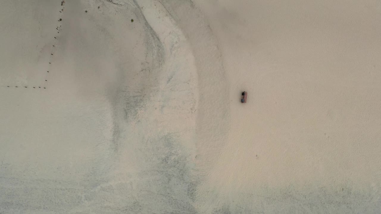 Overhead Los Cabos beach view showing shallow water along the left side, broad sand, a small dark object on the beach, and no visible people.