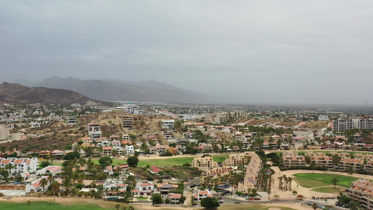 Opening aerial frame of Los Cabos showing hillside homes, low-rise buildings, roads, green grounds, distant mountains, and no visible people.
