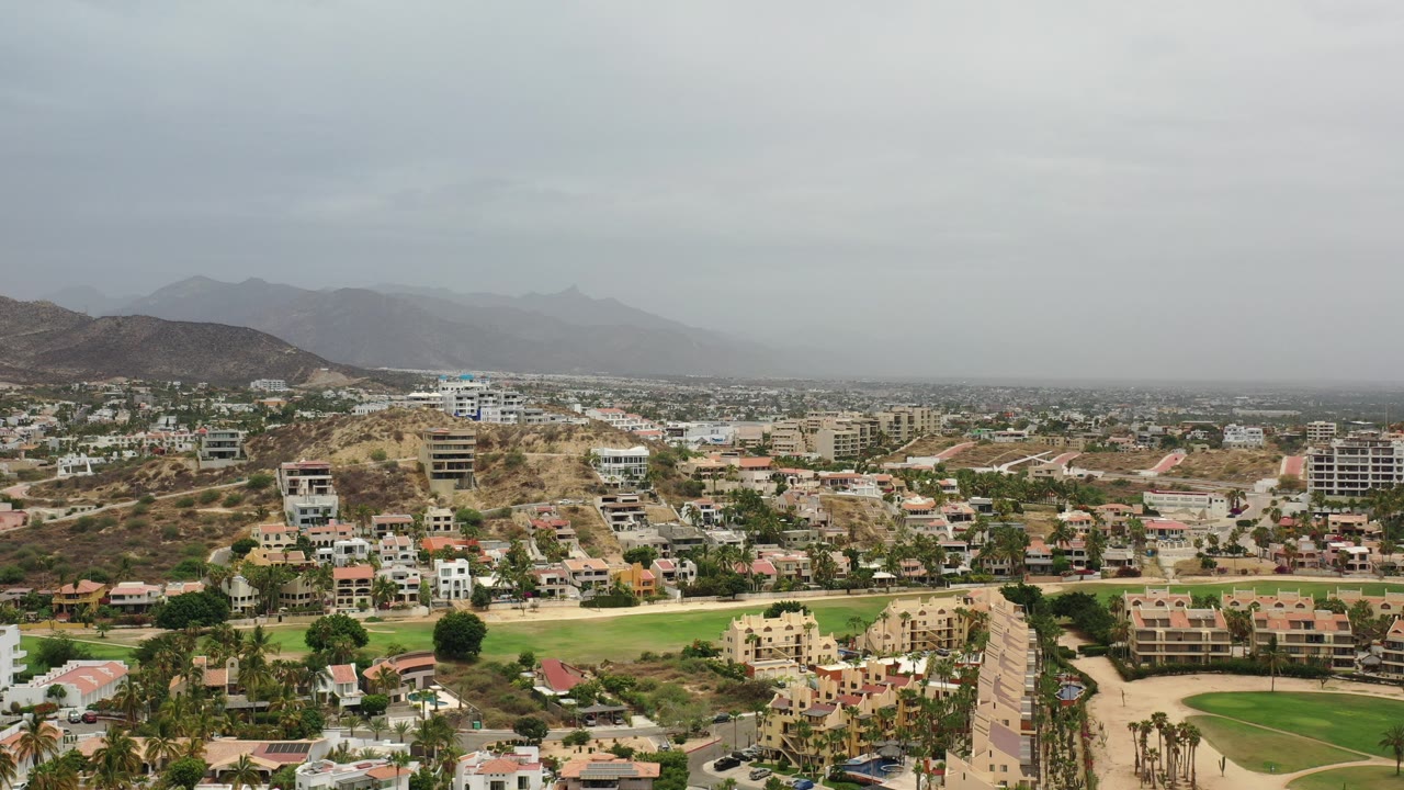 Aerial view of hillside buildings, golf course, roads, and distant mountains under gray clouds.