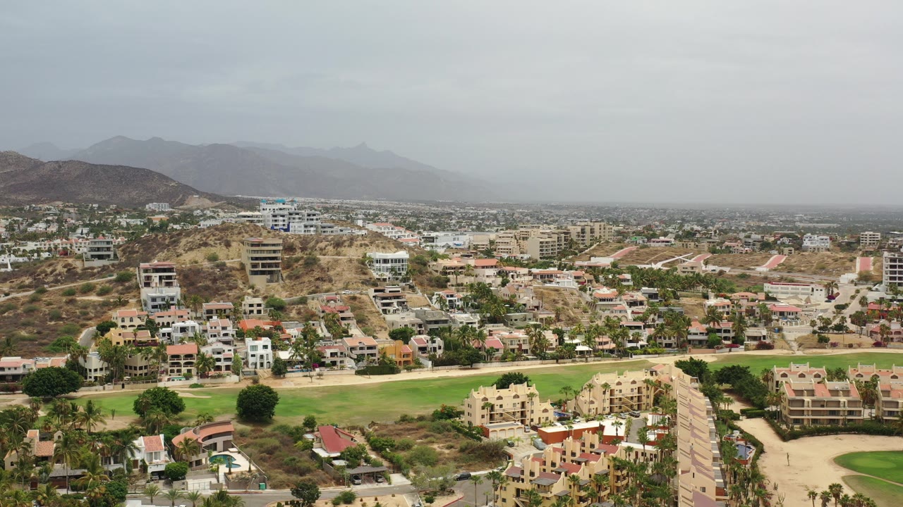 Wide elevated view over Los Cabos, Mexico, showing hillside homes, a broad green lawn, low-rise buildings, and hazy mountains in the distance, with no visible people.