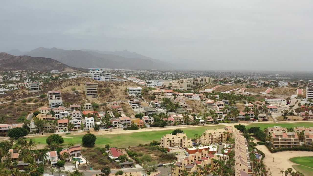 Elevated Los Cabos, Mexico view with modern houses clustered below a dry hillside, green grounds across the lower frame, and a hazy mountain horizon, with no visible people.
