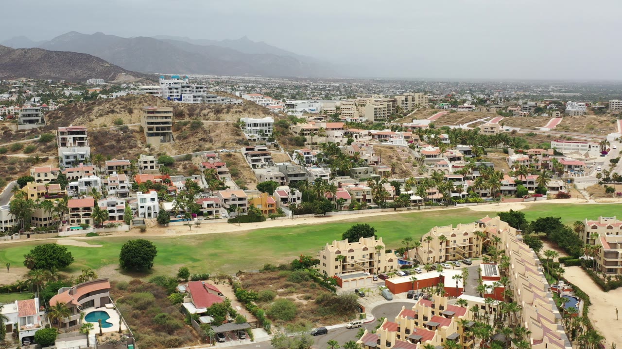 Wide aerial view in Los Cabos, Mexico, with a green field across the lower frame, residential buildings on the hillside, distant city blocks, and hazy mountains beyond.