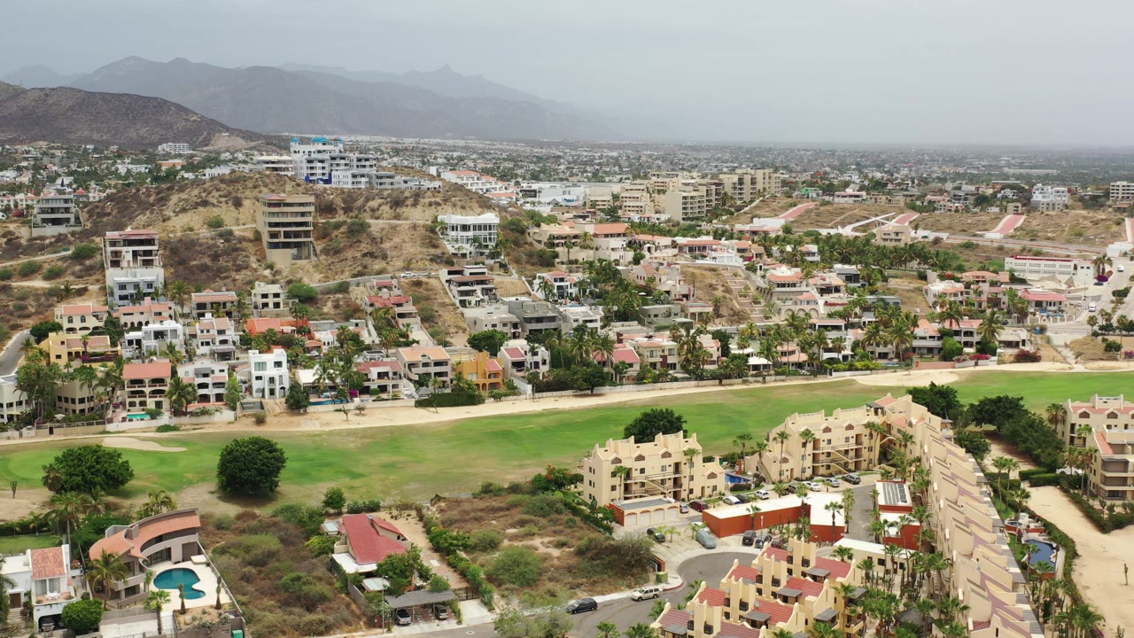 Los Cabos aerial frame showing a broad green strip in front of beige residential blocks, scattered trees, hillside homes, and a pale mountain horizon.